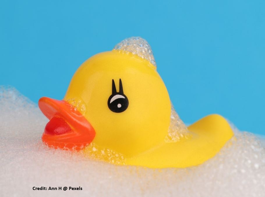 A smiling yellow Rubber Duck with painted eyelashes - probably manufactured using a plasticiser - languishes in soap suds. 